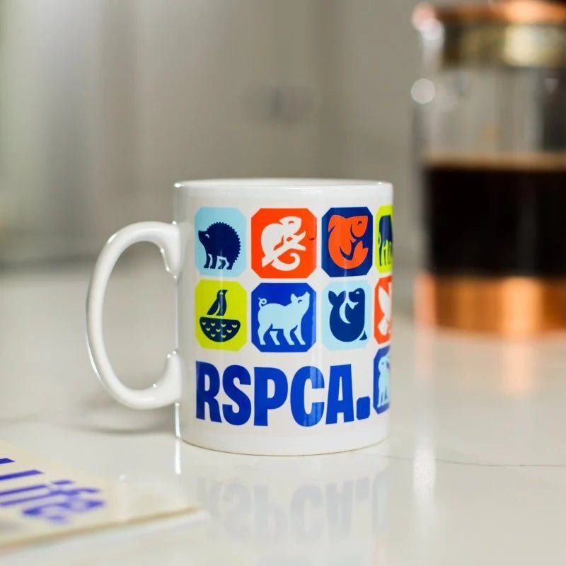 A white mug embossed with different animal graphics and the RSPCA logo. The mug is placed on a kitchen table with a cafetiere in the background. 