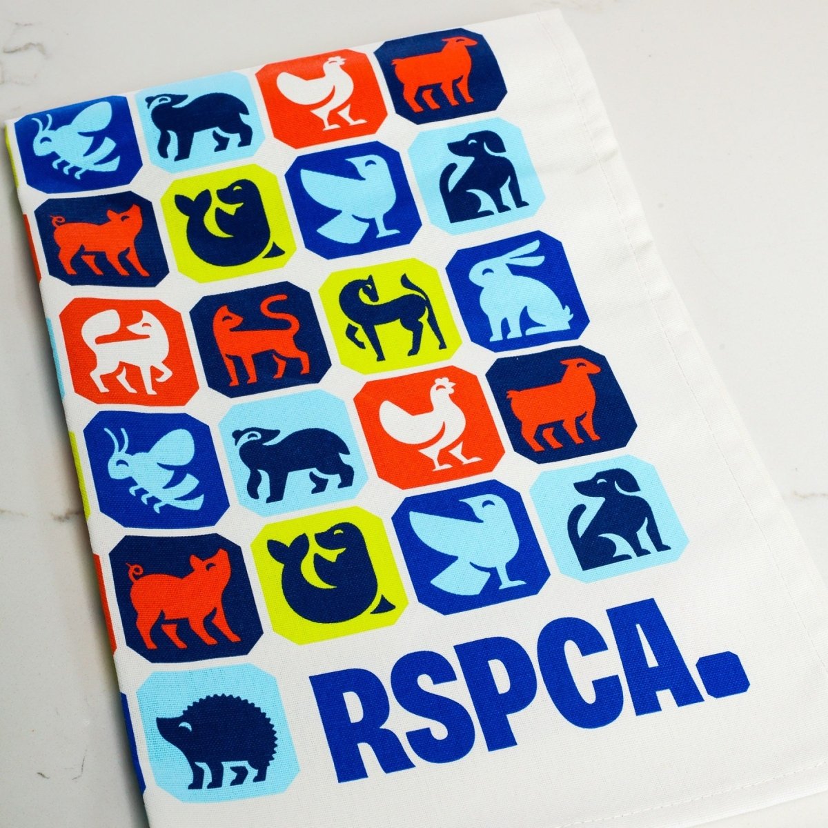 A folded RSPCA tea towel printed with different animal graphics and a blue RSPCA logo.