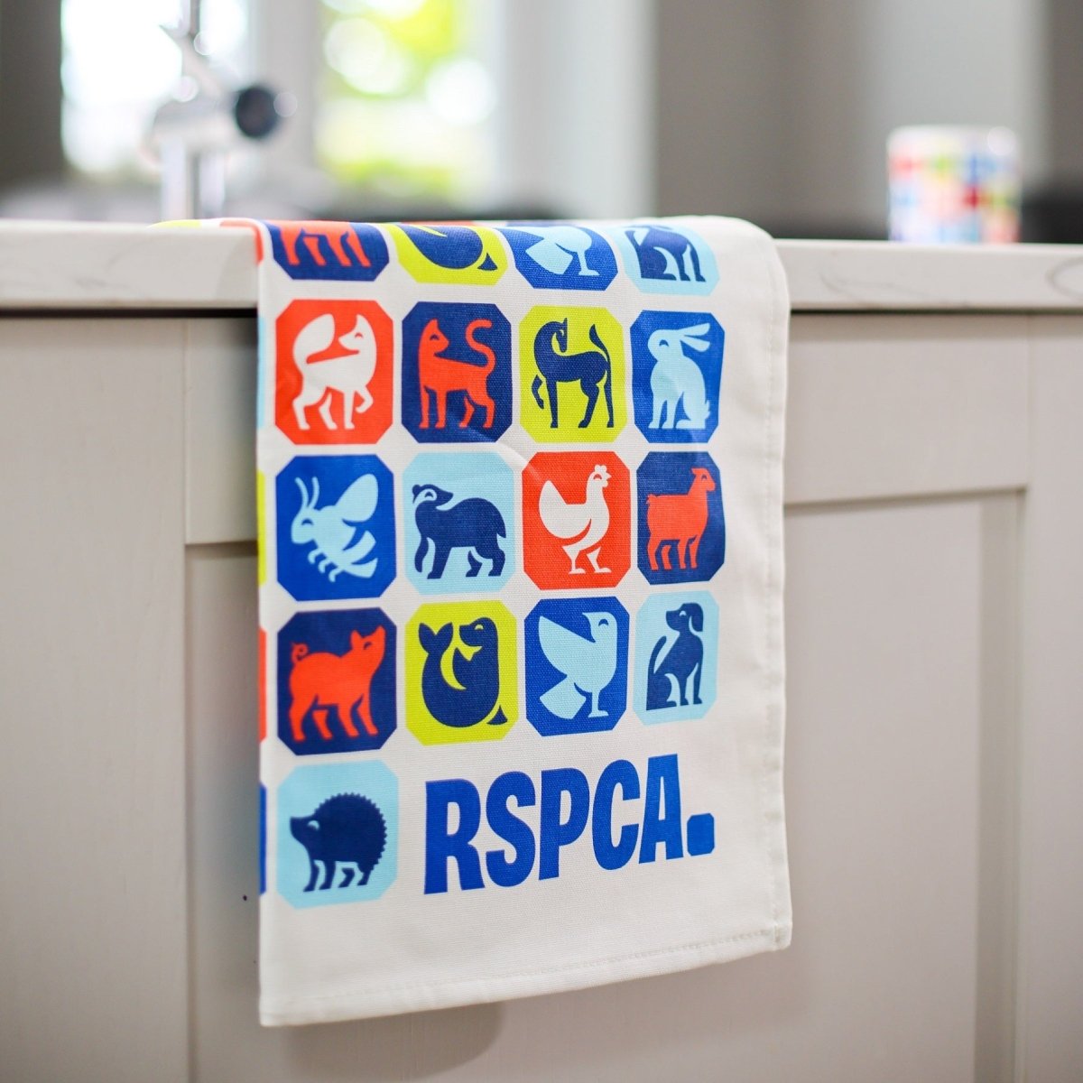 An RSPCA tea towel, printed with different animal graphics and a blue RSPCA logo, hanging over the side of a kitchen counter. 