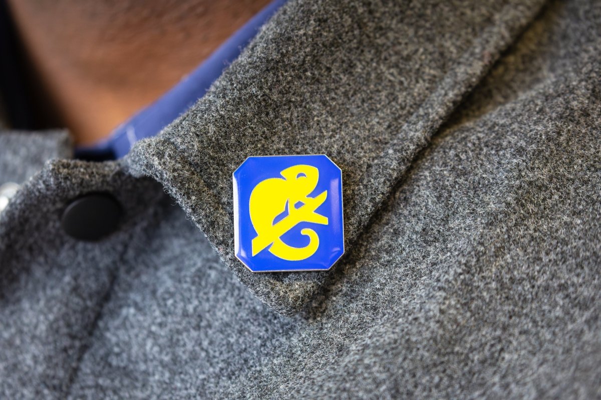 A metal pin badge embossed with a yellow chameleon graphic on a blue background, pinned to a grey coat collar. 