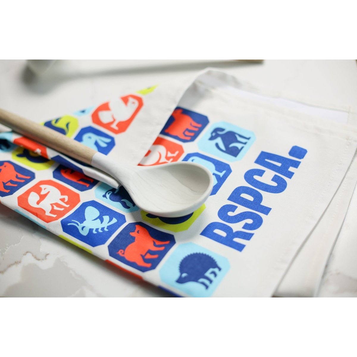 An RSPCA tea towel printed with different animal graphics and a blue RSPCA logo. A mixing spoon has been laid on top of the tea towel. 