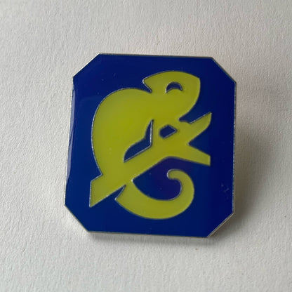 A blue metal pin badge embossed with a yellow chameleon graphic, against a white background. 