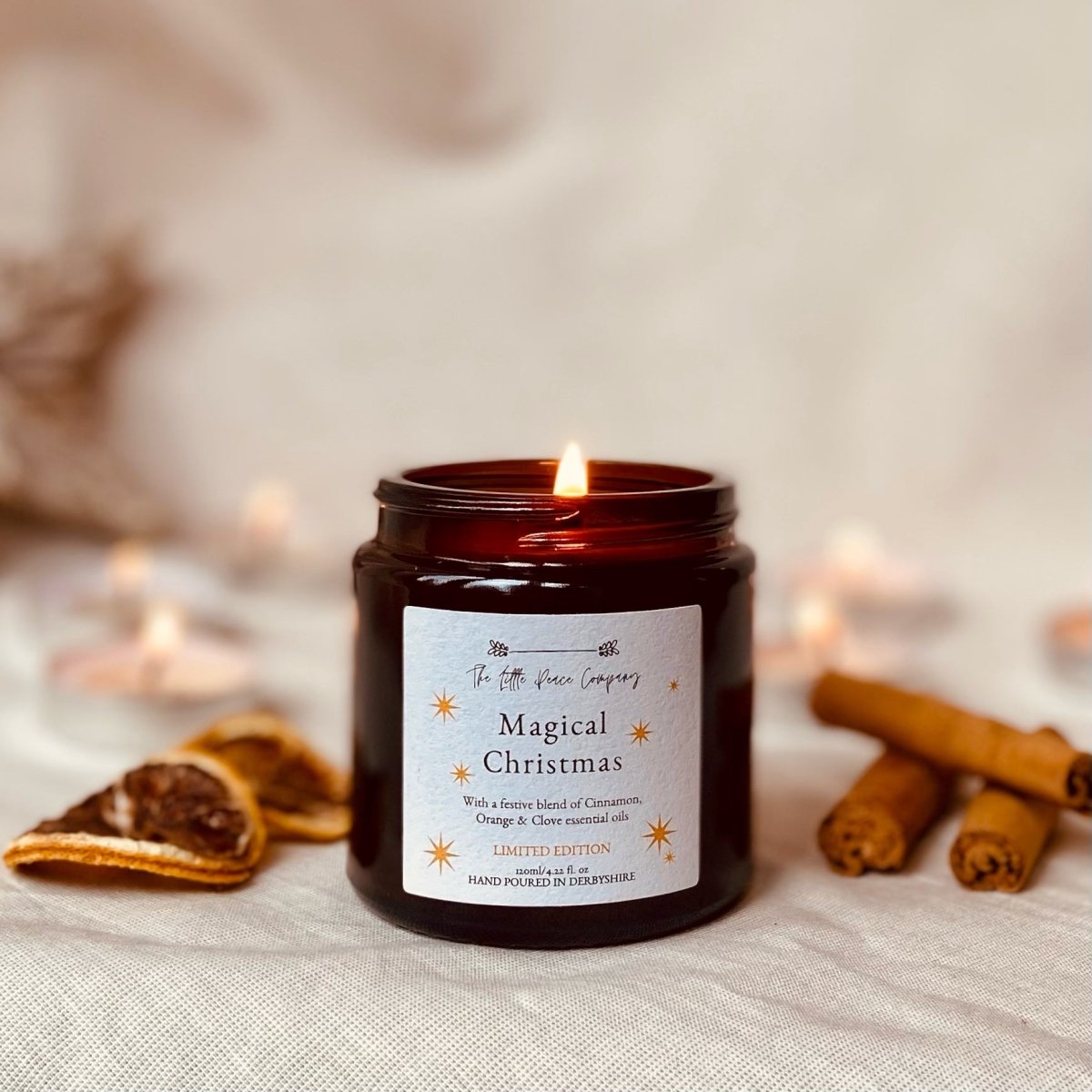 A Magical Christmas scented candle in a brown glass jar, dried orange slices and cinnamon sticks can be seen in the background. 