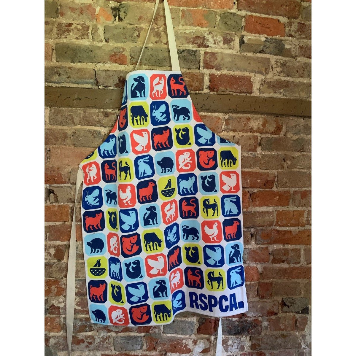 An apron printed with different animal graphics and the RSPCA logo on it. The apron is hanging on a hook against a brick wall. 