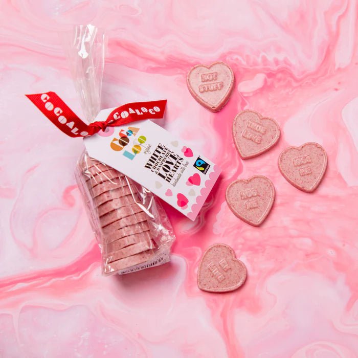 A pack of Cocoa Loco White Chocolate & Raspberry Love Hearts against a pink marble background. The packaging features a Fairtrade logo.